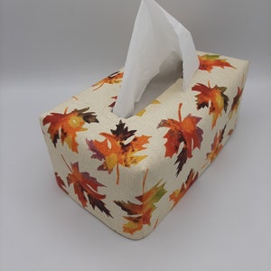 May include: A rectangular tissue box cover with a beige background and a repeating pattern of colorful autumn leaves in shades of orange, red, and yellow. White tissues are emerging from the top opening. The cover is made of a textured fabric.