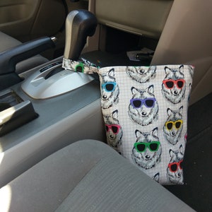 May include: A fabric car trash bag featuring a repeating pattern of wolves wearing sunglasses in various colors. The bag is attached to the gear shift. The background shows the interior of a car.
