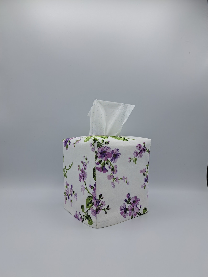 Puede incluir: Una funda cuadrada para caja de pa&ntilde;uelos con fondo blanco y un estampado floral de flores moradas y hojas verdes. Los pa&ntilde;uelos blancos sobresalen de la abertura superior. La funda es de tela.