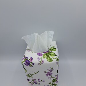 Puede incluir: Funda de caja de pa&ntilde;uelos cuadrada con un estampado floral blanco con flores moradas y hojas verdes. Los pa&ntilde;uelos blancos son visibles en la parte superior. Funda de tela.