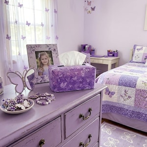 May include: A lavender bedroom with butterfly decor. A dresser with a butterfly-covered tissue box, a butterfly mirror, and a framed photo. A bed with a patchwork quilt and a butterfly rug complete the room's theme.