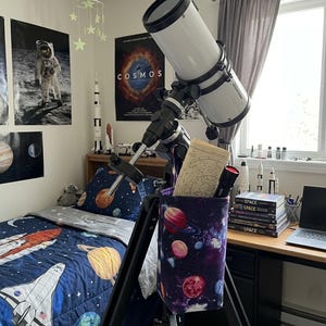 May include: A white telescope with a black tripod is set up in a bedroom. The room has space-themed decor, including posters, bedding, and a storage bag with planets. A red flashlight and star chart are in the telescope's storage bag.