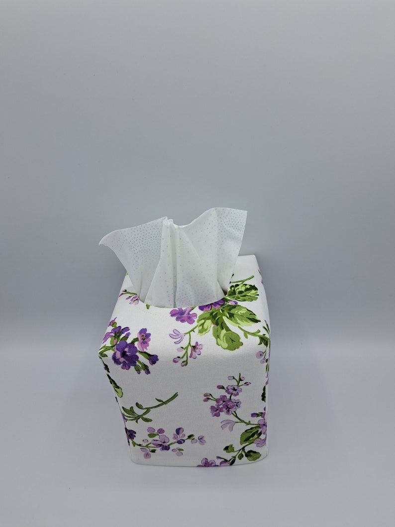 Puede incluir: Una funda blanca para caja de pa&ntilde;uelos con un estampado floral de flores moradas y hojas verdes. Los pa&ntilde;uelos son visibles en la parte superior. La caja tiene forma cuadrada.