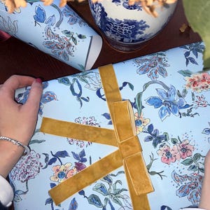 May include: A light blue gift box with a floral pattern of blue, pink, and peach flowers, tied with a gold velvet ribbon. A matching floral-patterned container is visible in the background, along with a blue and white ceramic vase.