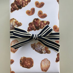 May include: White gift wrap with a navy blue and white striped ribbon. The wrapping paper features a repeating pattern of brown pastries, including croissants, madeleines, and palmiers.