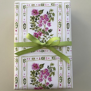 May include: A white gift box with a green ribbon tied in a bow. The box is wrapped in paper with a floral pattern of pink flowers and green leaves. The pattern is framed by a white border with a green and white geometric design.