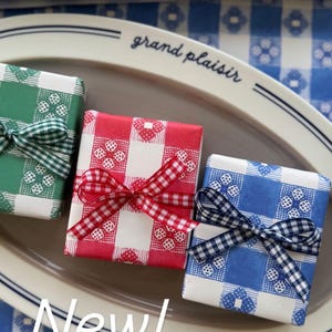May include: Three wrapped gifts in green, red, and blue picnic check paper, each tied with a matching ribbon. The gifts are on a gray oval platter, with a blue and white checkered background. Text on the image reads "New! Picnic Check Wrap."