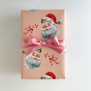 May include: A pink gift box with a pink satin bow. The box is wrapped in paper with a pattern of Santa Claus faces and candy canes.