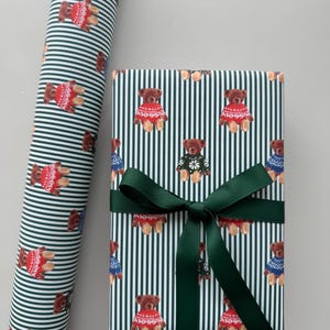 May include: Gift wrap featuring a repeating pattern of brown teddy bears wearing sweaters on a striped background. The wrapped gift is secured with a dark green ribbon. A roll of matching wrapping paper is visible.