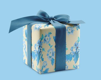 May include: A square gift box wrapped in cream-colored paper with a blue floral pattern. The box is tied with a wide, dark blue ribbon and a matching bow. The background is a solid light blue.