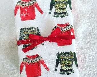 Può includere: Carta da regalo bianca con un motivo ripetuto di maglioni colorati. I maglioni sono rossi, verdi e bianchi con un motivo a intarsio. Un nastro rosso è legato attorno alla carta.
