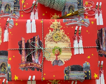 May include: A wrapped gift on red wrapping paper with a repeating pattern of cityscapes, soldiers, and stars. The gift is tied with green and white twine. A pair of scissors rests on the wrapping paper next to a spool of twine.