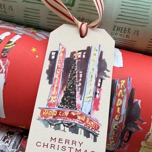 May include: Gift tags featuring a watercolor illustration of Radio City Music Hall with a Christmas tree. The tags have a cream background with red and gold accents, and the text "Merry Christmas" is printed on them. A red ribbon is attached.