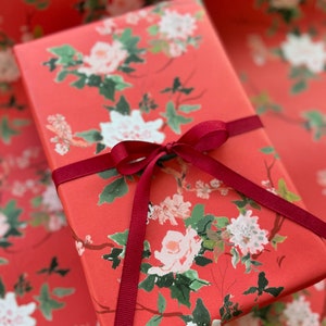 May include: A wrapped gift box featuring a floral pattern of white and pink flowers with green leaves on a red background. The box is tied with a burgundy ribbon, creating a festive and elegant presentation. The wrapping paper has a repeating floral design.