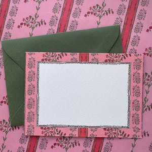 May include: A rectangular note card with a white centre and a pink border featuring a floral pattern and a hand-drawn black scalloped edge. The card is placed on top of a solid olive green envelope, both set against a pink floral background.