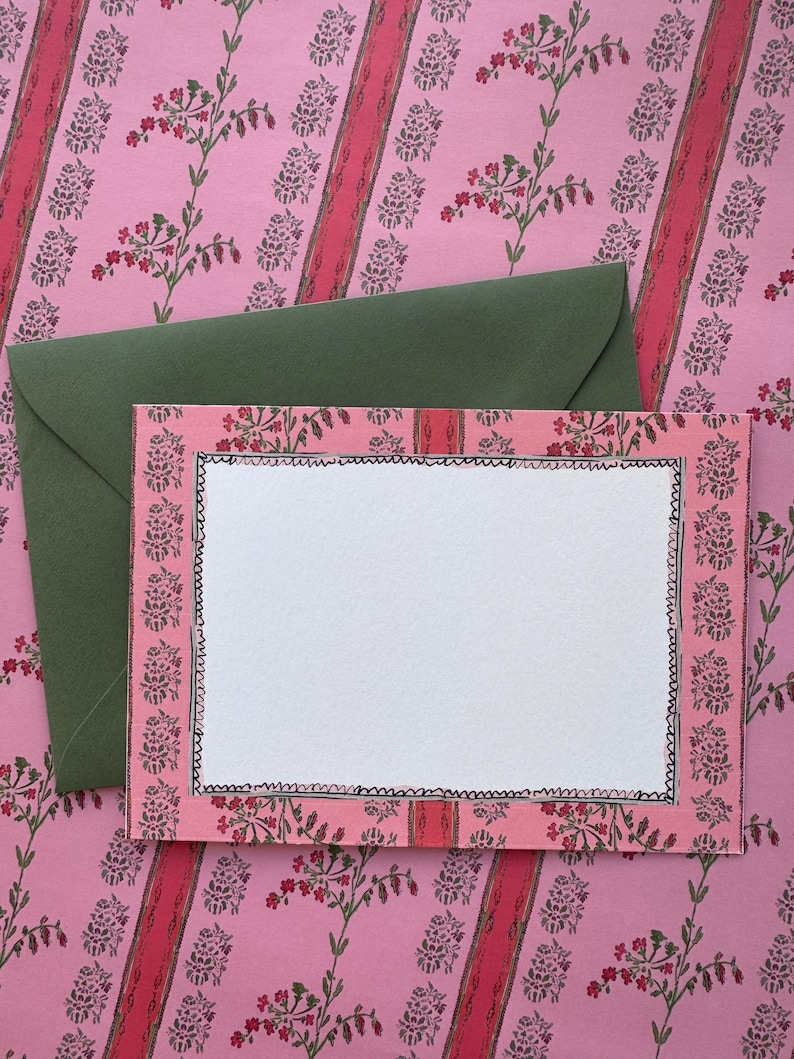 May include: A rectangular note card with a white centre and a pink border featuring a floral pattern and a hand-drawn black scalloped edge. The card is placed on top of a solid olive green envelope, both set against a pink floral background.