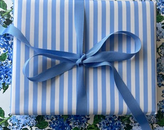 May include: A wrapped gift with blue and white striped paper, tied with a light blue ribbon. The gift is on a background of blue hydrangea flowers.