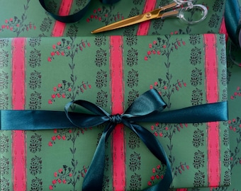 May include: A rectangular gift wrapped in green paper featuring a repeating pattern of dark green floral motifs and vertical bright pink stripes. A dark green satin ribbon is tied in a bow around the package, accompanied by gold-toned scissors.