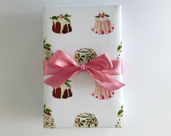 May include: White gift wrap with a pink ribbon and a repeating pattern of watercolor illustrations of Christmas pudding and cake.