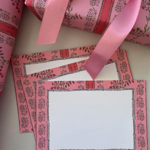 May include: A stack of rectangular note cards and a roll of wrapping paper, both featuring a pink background with a repeating pattern of green floral sprigs and vertical red stripes. A smooth pink satin ribbon rests across the stationery.