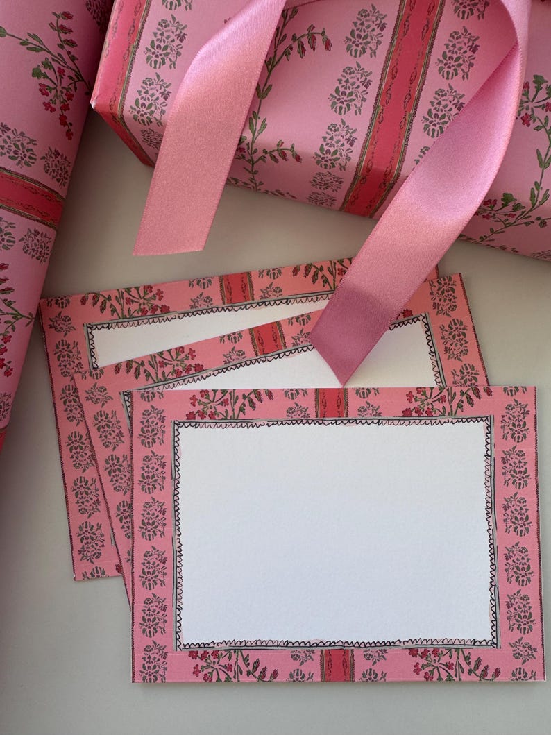 May include: A stack of rectangular note cards and a roll of wrapping paper, both featuring a pink background with a repeating pattern of green floral sprigs and vertical red stripes. A smooth pink satin ribbon rests across the stationery.