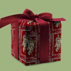 May include: A wrapped gift box with a red floral pattern and a matching burgundy ribbon and bow. The box is square and the ribbon is wrapped around the box. The background is a solid green color.