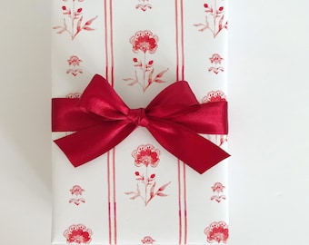 May include: A white gift box with a red ribbon and a floral pattern. The flowers are red and pink and have a watercolor look. The box is wrapped in white paper with red stripes.