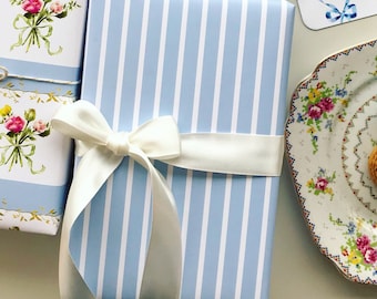 May include: A blue and white striped gift wrap with a white satin ribbon tied in a bow. The ribbon is tied in a bow on top of the gift wrap.