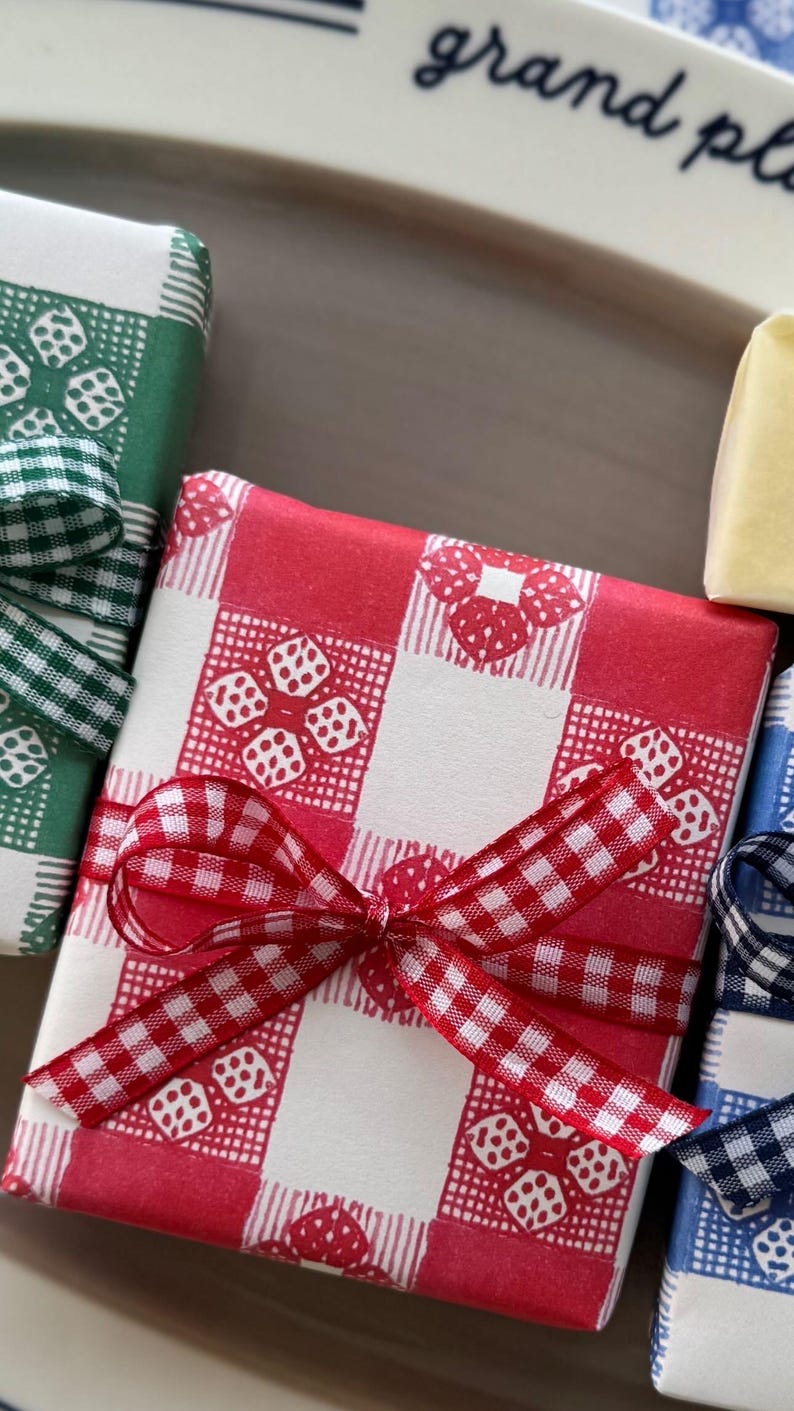 May include: Close-up of wrapped gifts with red, green, and blue gingham patterns. Each gift is tied with a matching gingham ribbon. The gifts are on a white plate with the words "grand plat" in cursive.