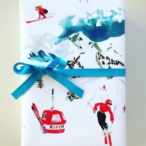 May include: White gift wrap with a blue ribbon. The wrap features a watercolor design of a snowy mountain scene with skiers and a ski lift. The ski lift has a red gondola with the word "ASPEN" printed on it.