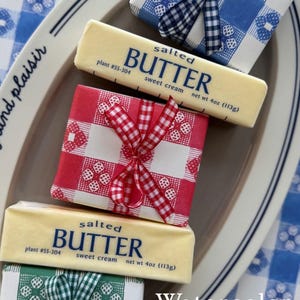 May include: Butter wrapped in watercolor picnic-themed paper. The butter is wrapped in red, blue, and green gingham patterns, tied with matching ribbons. The image includes the text "Watercolor Picnic Wrap".