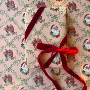 May include: A rectangular gift box wrapped in light pink paper featuring a repeating pattern of Santa Claus faces and silver bells with red bows, tied with a deep red velvet ribbon that forms a bow on the front surface of the package.