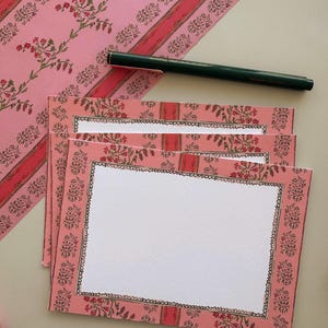 May include: A stack of rectangular note cards featuring a pink border with a repeating floral pattern in red and green, surrounding a white centre. A dark green pen rests diagonally across the top card, which has a black scalloped border.