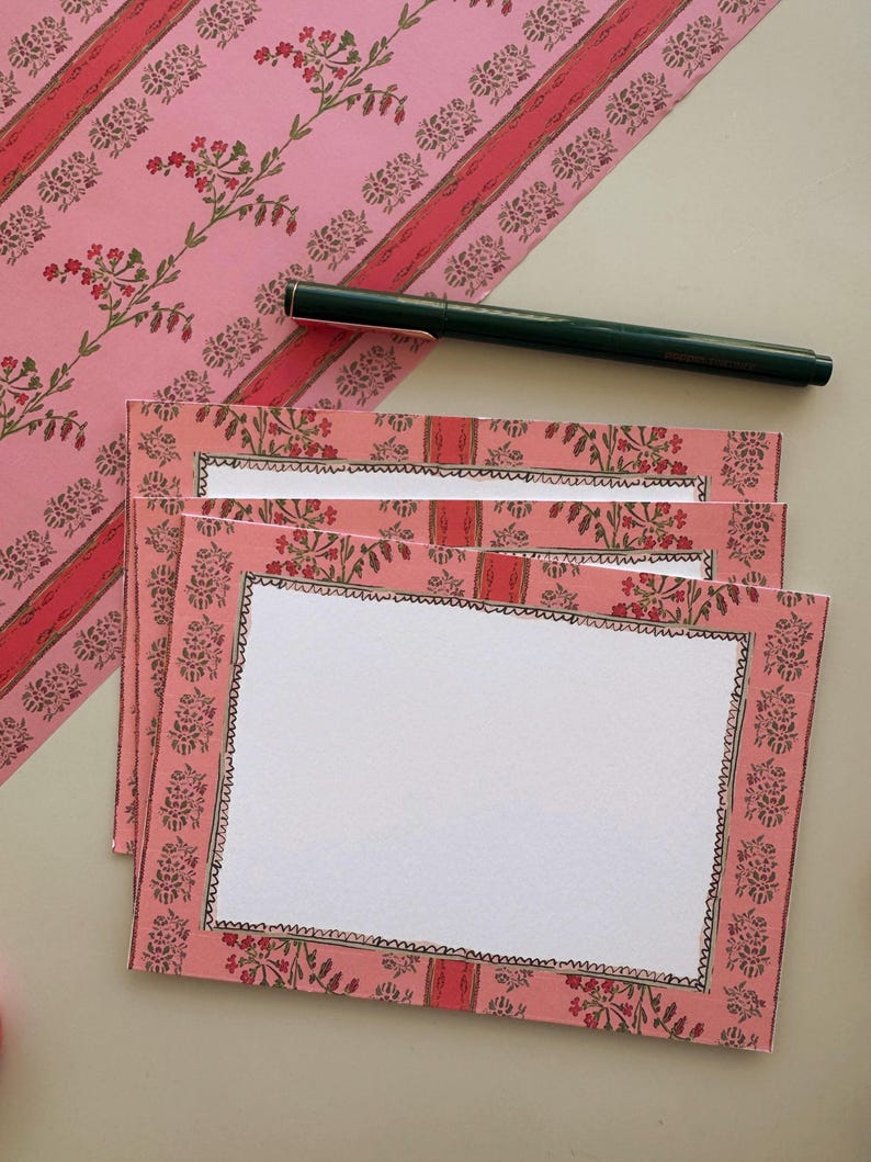 May include: A stack of rectangular note cards featuring a pink border with a repeating floral pattern in red and green, surrounding a white centre. A dark green pen rests diagonally across the top card, which has a black scalloped border.