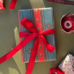 May include: A rectangular gift box wrapped in blue paper with vertical stripes and a central red floral band, tied with a bright red satin ribbon. The box is positioned on a flat surface alongside a roll of red ribbon and another gift.