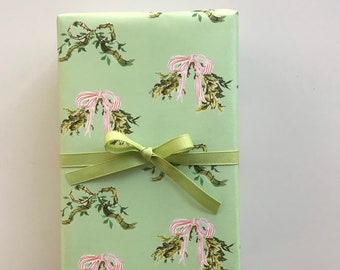 Puede incluir: Una caja de regalo rectangular envuelta en papel verde claro con un patrón repetido de coronas verdes y marrones con lazos rosas. Una cinta verde claro está atada alrededor de la caja.