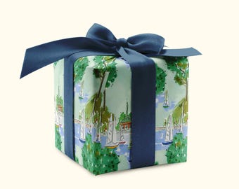 May include: A wrapped gift box with a navy blue ribbon and bow. The wrapping paper features a scenic illustration of sailboats on a lake with green trees and buildings. The box is square and the ribbon is tied around it.