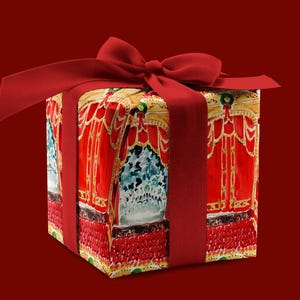 May include: A red gift box with a red ribbon and bow. The box is wrapped in paper with a colorful design of a stage curtain with gold trim.