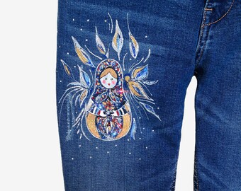 May include: Blue denim jeans with a hand-painted design of a Russian nesting doll, or matryoshka doll, with a floral pattern. The doll is surrounded by silver and gold glitter.