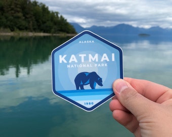 May include: A blue and white hexagonal sticker with the text "Katmai National Park" and "Alaska" in white. A blue bear silhouette is in the center of the sticker. The text "1980" is at the bottom of the sticker.