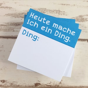 Può includere: Una pila di blocchi per appunti bianchi con un foglio superiore blu. Il foglio blu ha il testo bianco "Heute mache ich ein Ding." e la parola "Ding:" in blu. I blocchi per appunti sono su una superficie di legno chiaro.