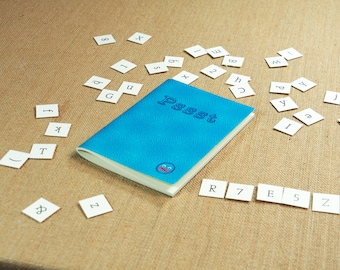 May include: A blue notebook with the word "Pssst" written on the cover. The notebook is surrounded by small white squares with letters and numbers on them.