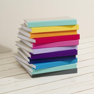Könnte beinhalten: Ein Stapel aus elf Hardcover-Büchern mit einfarbigen Stoffeinbänden in Hellblau, Gelb, Orange, Magenta, Pink, Lavendel, Violett, Dunkelblau, Cyan, Mintgrün und Dunkelgrau, die alle über weiße Buchseiten verfügen.