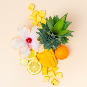 May include: A vibrant arrangement of tropical fruits and a flower on a light beige background. The image features a pineapple top, mango slices, orange, lemon and lime slices, and diced pineapple. A white hibiscus flower with a red center is also present.