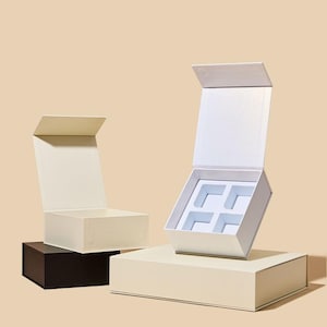 May include: Three open gift boxes in cream, white, and black. The white box has a light blue interior with four square compartments. The boxes are arranged on a cream-colored surface against a neutral background.