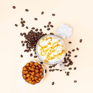 May include: Overhead shot of a clear glass mug filled with a coffee drink, topped with whipped cream and caramel drizzle. Scattered around are coffee beans, a small bowl of hazelnuts, and a dollop of whipped cream on a light beige background.
