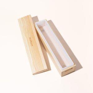 May include: A wooden soap mold with a removable lid. The mold is rectangular with a light-colored wood finish and a white silicone liner. The lid has the word "makeky" printed on it.