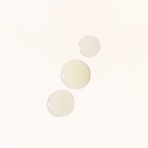 May include: Three light yellow liquid droplets against a white background. The droplets vary in size, with the largest at the bottom and the smallest at the top. The droplets appear smooth and reflective.