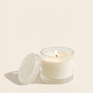 May include: A lit candle in a clear glass jar with a decorative lid. The candle has a white wax and a small flame. The jar and lid have a textured, geometric design. The background is a solid, light beige color.