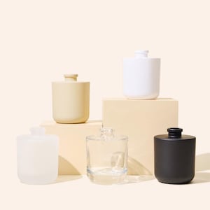 May include: A collection of ceramic and glass bottles in various colors, including white, cream, clear, and black. The bottles have a cylindrical shape with a rounded top and a small, raised neck. Some bottles are opaque, while others are transparent.
